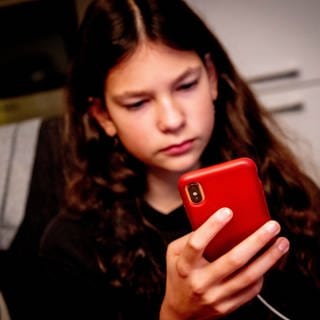 Ein Mädchen schaut ernst auf sein rotes Smartphone: Immer mehr Teenager verlieren sich in der digitalen Welt und kämpfen mit der Realität, einige entwickeln Suchtverhalten, Ängste oder Depressionen. Medizin wie Wissenschaft entwickeln für die wachsende Zahl Betroffener kreative Therapien und präventive Ansätze und holen die Eltern mit ins Boot.