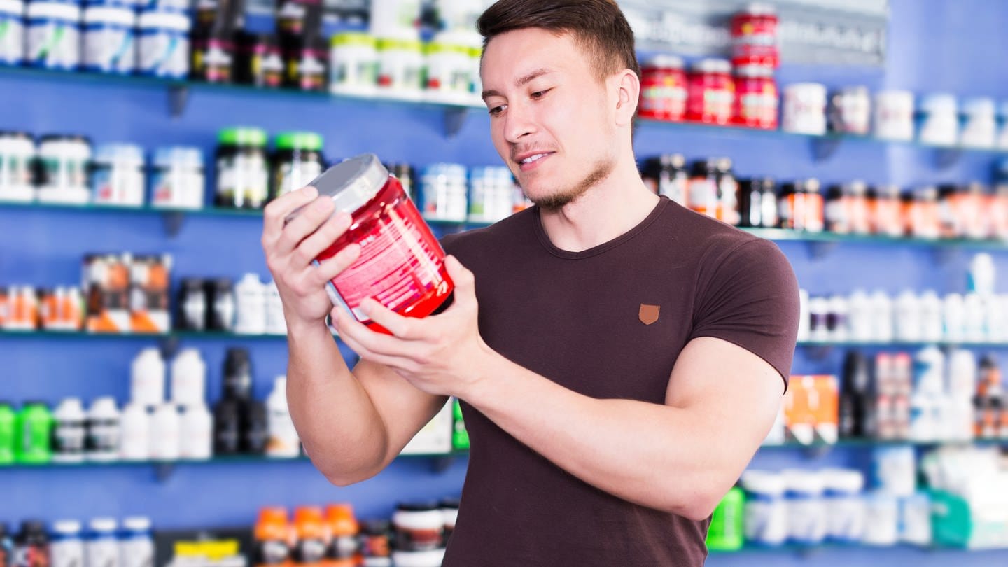 Ein junger Mann liest die Inhaltsangabe eines Protein-Shakes in einem Laden; im Hintergrund viele weitere Proteinprodukte: Viele sportlich aktive Menschen möchten ihre Fitness durch Ernährung unterstützen, wobei Proteine eine wichtige Rolle spielen. Die Industrie hat hier einen lukrativen Markt entdeckt.