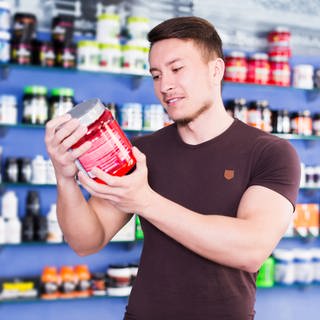 Ein junger Mann liest die Inhaltsangabe eines Protein-Shakes in einem Laden; im Hintergrund viele weitere Proteinprodukte: Viele sportlich aktive Menschen möchten ihre Fitness durch Ernährung unterstützen, wobei Proteine eine wichtige Rolle spielen. Die Industrie hat hier einen lukrativen Markt entdeckt. 