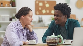 Junges Paar unterhält sich am Tisch bei einer Tasse Kaffee: Nicht ohne Grund wird in vielen Sprachen zwischen passivem Hören und aktivem Zuhören unterschieden. Welche Rolle spielt Empathie in der Kommunkation? Wie lässt sich gutes Zuhören erlernen? Und welche Auswirkungen kann das auf die Beteiligten haben?