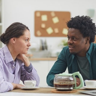 Junges Paar unterhält sich am Tisch bei einer Tasse Kaffee: Nicht ohne Grund wird in vielen Sprachen zwischen passivem Hören und aktivem Zuhören unterschieden. Welche Rolle spielt Empathie in der Kommunkation? Wie lässt sich gutes Zuhören erlernen? Und welche Auswirkungen kann das auf die Beteiligten haben?