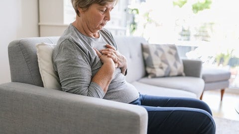 Eine Frau sitzt auf einem grauen Sofa und greift sich an die Brust: Der weibliche Herzinfarkt wird oft später erkannt und später behandelt. 