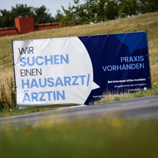 Ein Plakat mit dem Text "Wir suchen einen Hausarzt  Ärztin - Praxis Vorhanden" steht am Ortseingang von Friederichskoog. Hausärzte werden in vielen ländlichen Regionen gesucht.