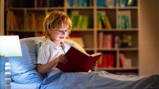 Ein Kind liest abends im Bett in einem großen Buch: Viele Kinder lesen nicht gern – selbst, wenn ihnen zu Hause regelmäßig vorgelesen wurde. Zwar steigt dadurch eigentlich die Wahrscheinlichkeit, dass Kinder später selbst gern zum Buch oder E-Book greifen, ein Garant ist es aber nicht. 