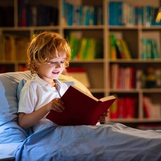 Ein Kind liest abends im Bett in einem großen Buch: Viele Kinder lesen nicht gern – selbst, wenn ihnen zu Hause regelmäßig vorgelesen wurde. Zwar steigt dadurch eigentlich die Wahrscheinlichkeit, dass Kinder später selbst gern zum Buch oder E-Book greifen, ein Garant ist es aber nicht. 