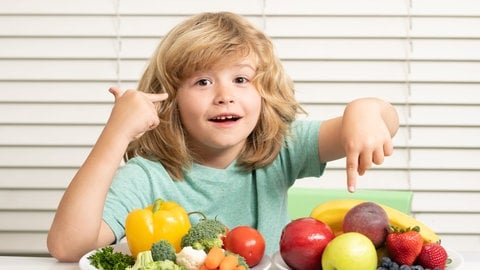 Kind sitzt vor zwei Tellern mit Gemüse und Obst: Kinder sollten schon früh an gesundes Essen gewöhnt und an Bewegung herangeführt werden. Aber zu der Frage, was Kinder tatsächlich selbst über eine gesunde Lebensweise wissen, ist die Studienlage dünn.