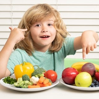 Kind sitzt vor zwei Tellern mit Gemüse und Obst: Kinder sollten schon früh an gesundes Essen gewöhnt und an Bewegung herangeführt werden. Aber zu der Frage, was Kinder tatsächlich selbst über eine gesunde Lebensweise wissen, ist die Studienlage dünn.