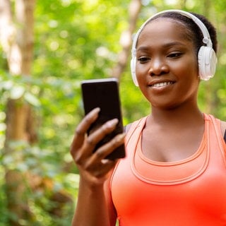 Junge Frau mit Kopfhörer und Smartphone geht durch den Wald: Das Radio der Zukunft soll die Hörerinnen und Hörer unterwegs mit relevanten Informationen versorgen und mittels KI mit ihnen in Dialog treten können