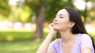 Junge Frau sitzt zufrieden lächelnd mit geschlossenen Augen im Park: Zeit mit sich allein zu verbringen ist entscheidend, um sich selbst zu verstehen, auszuprobieren und innerlich zu wachsen. Psychologinnen und Therapeuten halten Alleinsein für eine bedeutende Fähigkeit. Sie kann davor schützen, in Abhängigkeiten zu geraten oder in Einsamkeit. 