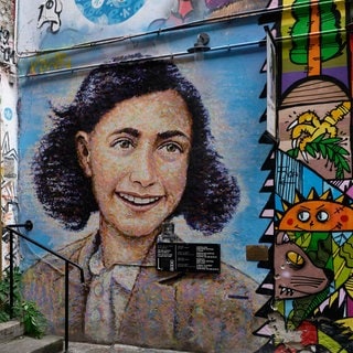 Porträt von Anne Frank als Mauerbild am Anne-Frank-Zentrum in den Hackeschen Höfen in Berlin: Anne Franks Tagebücher von 1942 bis 1944 gelten als eines der bekanntesten persönlichen Zeugnisse der NS-Zeit. Das hat zu einem globalen Personenkult geführt. Während die wissenschaftliche Aufarbeitung der Tagebücher noch nicht abgeschlossen ist und die Umstände von Anne Franks Verhaftung und Tod rätselhaft bleiben, überlagert der Kult die Erinnerung an den Holocaust. 