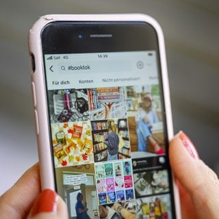 Zwei Hände mir rot lackierten Fingernägeln halten ein Smartphone, auf dem in einer Social-Media-App der Filter #booktook aktiviert ist: Booktoker und Bookfluencer besprechen auf Insta und Tiktok Bücher. Ihre Follower feiern sie dafür wie Popstars. Das bringt Verlagen neue Umsätze und mischt den Literaturbetrieb auf. 