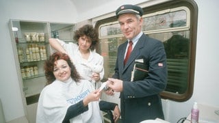 Intercity-Friseur im Mai 1989. Eine Frau bekommt die Haare frisiert, ein Zugbegleiter kontrolloiert den Fahrschein: 