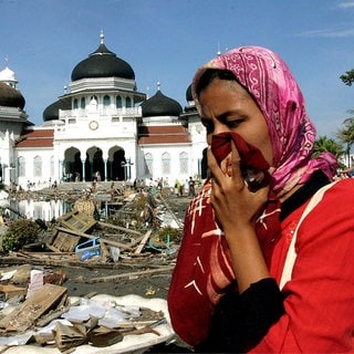 Eine Frau bedeckt ihre Nase mit einem Tuch; im Hintergrund eine Moschee. In den Straßen ist der Müll zu sehen, der sich nach dem Erdbeben und Tsunami in Banda Aceh auf der indonesischen Insel Sumatra im Dezember 2004 angesammelt hat.