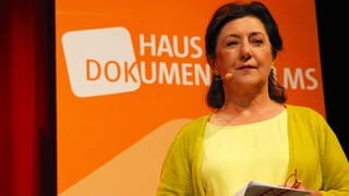 Die Geschäftsführerin im Haus des Dokumentarfilms, Ulrike Becker, steht vor einer Logowand und trägt ein Mikrofon