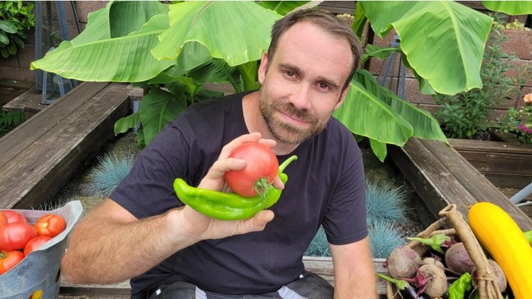 Miguel ist in seinem Selbstversorgergarten, mit einem Teil seiner Ernte - rote Beete, Tomaten, Zucchini und Parpika. 