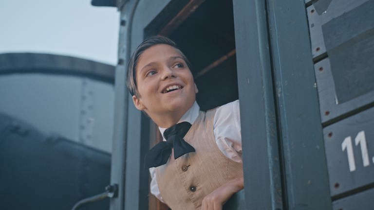 Thomas Alva Edison (Emile Cherif) beugt sich aus dem Fenster des fahrenden Zugs und lächelt.