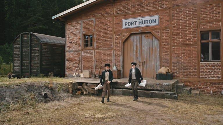 James (Ilja Bultmann, re.) und Alva (Emile Cherif, li.) laufen aus einem Gebäude mit dem Titel "Port Huron". In den Händen haben sie kleine Säckchen.