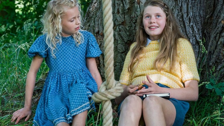 Judy (Selma Suckfüll, links) und Jane (Aurelia Ott, rechts), im Gras sitzend, angelehnt an einen Baum. Judy bringt Jane auf die eine Idee, wie sie das Pferd Wellington vorm Schlachter retten können.