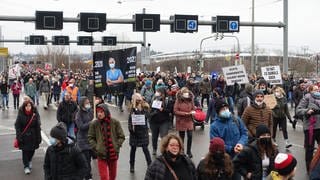 Viele gegen die Corona-Maßnahmen Protestierende laufen wegen der kalten Jahreszeit im Januar in dicke Jacken gehüllt und mit Mützen in einem Protestzug auf einer breiten Straße in Stuttgart. Einige tragen Schilder oder Plakate, manche mit, aber viele ohne Mund-Nasen-Schutz.