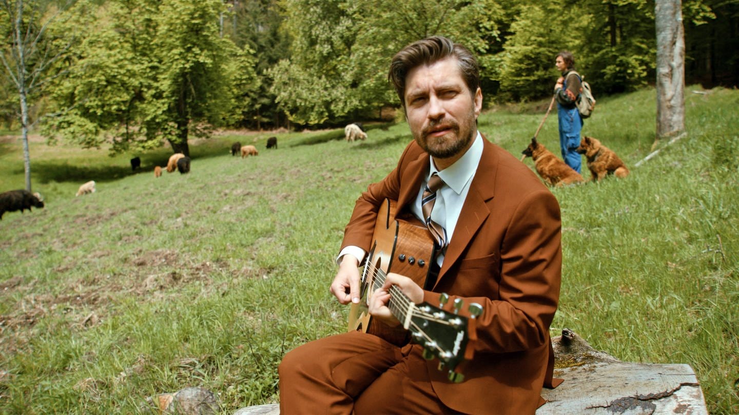 David Julian Kirchner spielt ein Lied auf der Gitarre inmitten einer Kuh-Herde