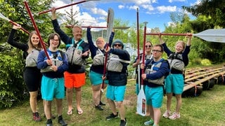 Die Reisegruppe hat ein Floß gebaut und paddelt damit über den Schluchsee. Alle haben eine Rettungsweste an und recken ein Paddel in die Höhe.