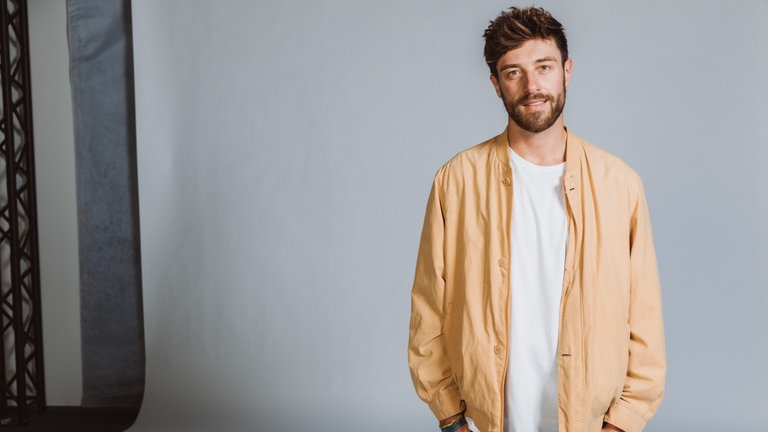 "Ausgesprochen Geil"-Host Frederik Fleig steht in einem gelben Blouson vor einer Fotoleinwand.