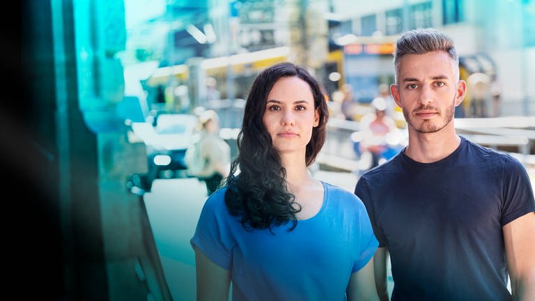 Leonie Maderstein (li.) und Marvin Neumann (re.) beim Fotoshooting in Stuttgart. Beide tragen blaue T-shirts. Im Hintergrund ist verschwommen eine Straßenbahn zu sehen.