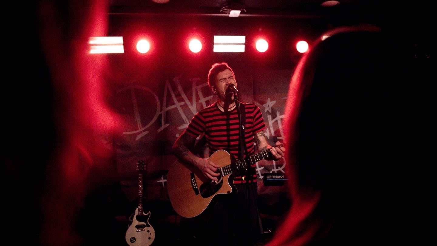 Dave im roten Scheinwerferlicht mit einer Gitarre singend auf der Bühne