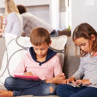 Ein Junge und ein Mädchen mit Smartphone und Tablet auf dem Sofa, im Hintergrund Erwachsene