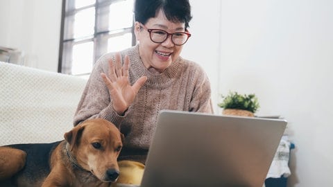 Ältere asiatische Dame mit Hund vor Laptop, sie winkt freundlich. Immer mehr Menschen nutzen Livestream-Shopping.