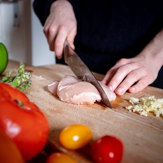 Eine Person schneidet ein Stück Fleisch auf einem Holz-Schneidebrett. Ein Labortest zeigt: Viele Schneidebretter in der Küche aus Holz oder Plastik sind mit Keimen belastet. Welches Brett sich wofür eignet und was man bei der Reinigung beachten sollte.