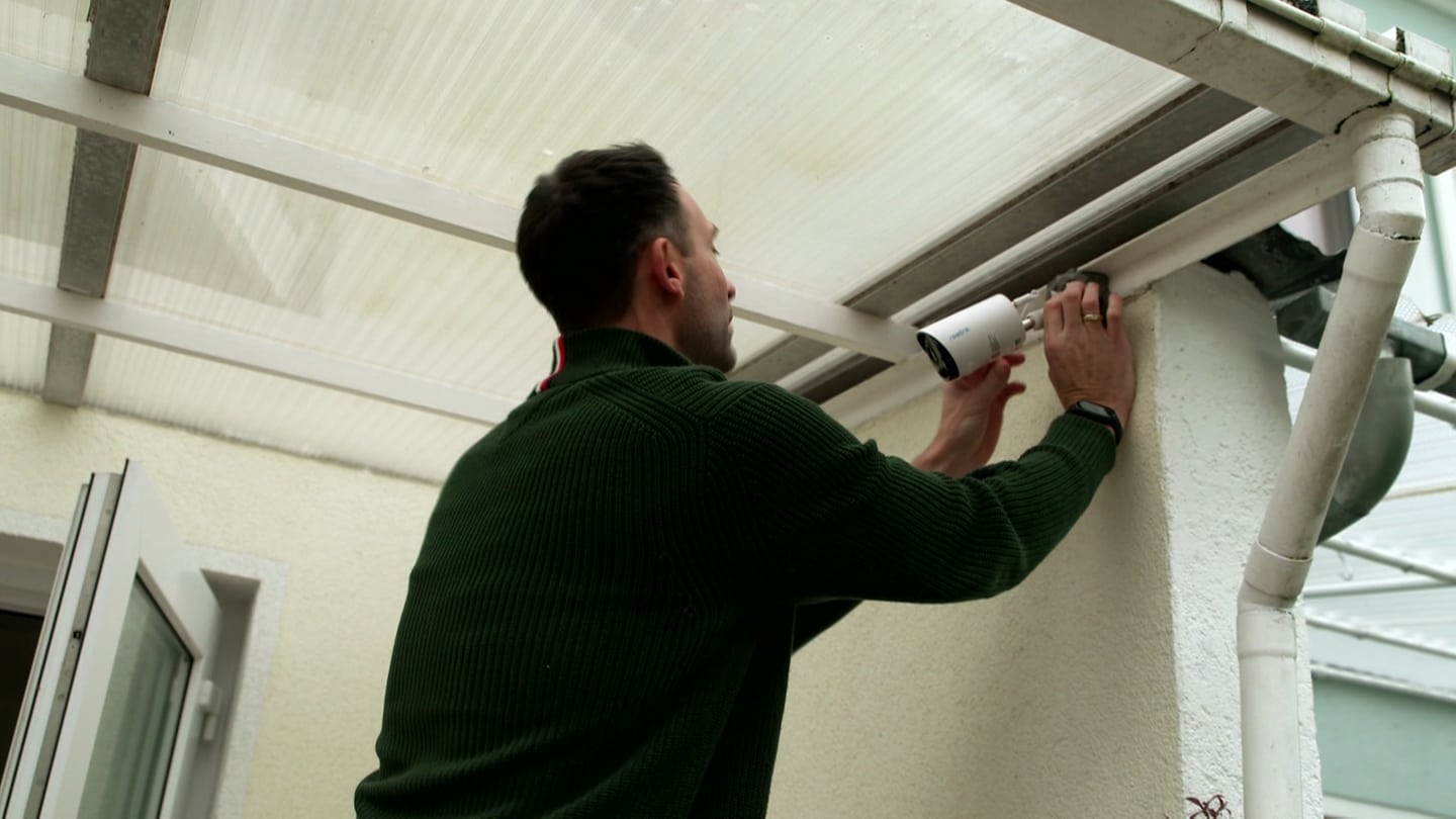 Ein Mann installiert eine Überwachungskamera an einer Hauswand oben. Vor einem Einbruch geschützt zu sein, wünscht sich jeder. Videokameras sollen das eigene Zuhause sichern und im Ernstfall Besitzer alarmieren. Doch es gibt Qualitätsunterschiede.