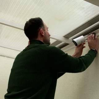 Ein Mann installiert eine Überwachungskamera an einer Hauswand oben. Vor einem Einbruch geschützt zu sein, wünscht sich jeder. Videokameras sollen das eigene Zuhause sichern und im Ernstfall Besitzer alarmieren. Doch es gibt Qualitätsunterschiede.