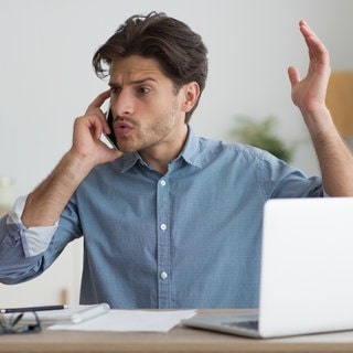 Junger Mann in blauem Hemd vor Laptop telefoniert und gestikuliert wütend am Handy. Was ist die neue Abhilfeklage? Wie können Sie gegen Vodafone, Facebook & Co. vorgehen?