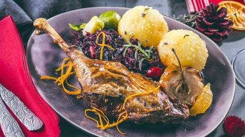 Gänsekeule mit Rotkohl und hellen Knödeln. Dazu eine dunkle Soße, ein halber Apfel aus dem Ofen und Rosenkohl. Garniert mit Rosmarin und Orangenzesten.