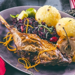 Gänsekeule mit Rotkohl und hellen Knödeln. Dazu eine dunkle Soße, ein halber Apfel aus dem Ofen und Rosenkohl. Garniert mit Rosmarin und Orangenzesten.