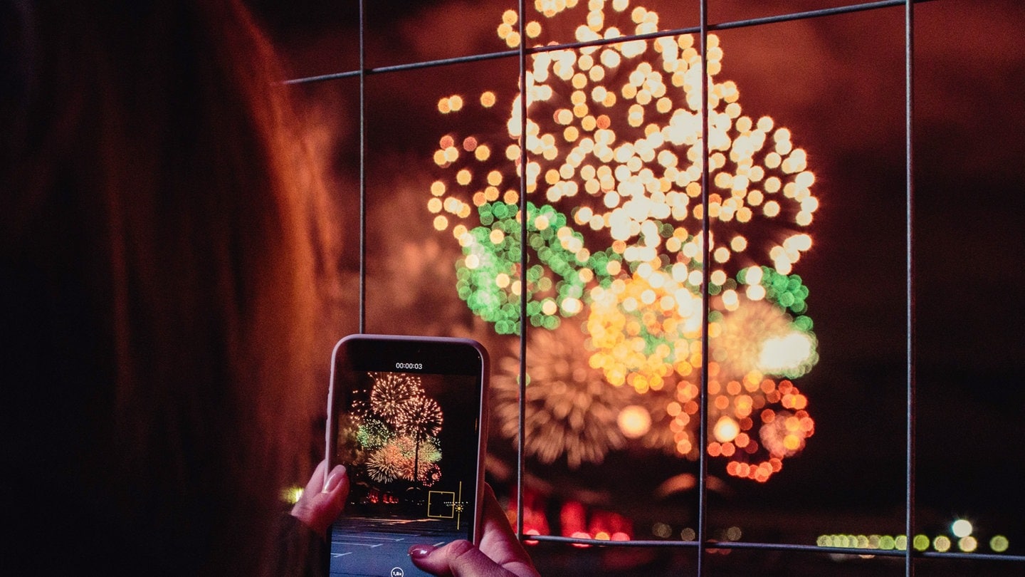 Frau fotografiert mir ihrem Smartphone ein Feuerwerk: Welche Versicherung zahlt Silvester Schäden durch Böller und Knaller an Autos, in Wohnungen oder auch bei Personen.