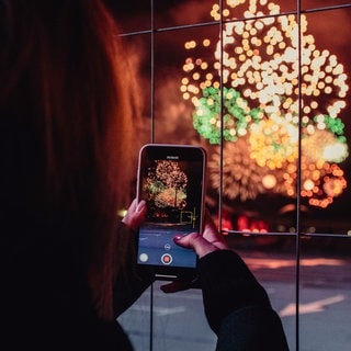 Frau fotografiert mir ihrem Smartphone ein Feuerwerk: Welche Versicherung zahlt Silvester Schäden durch Böller und Knaller an Autos, in Wohnungen oder auch bei Personen. 