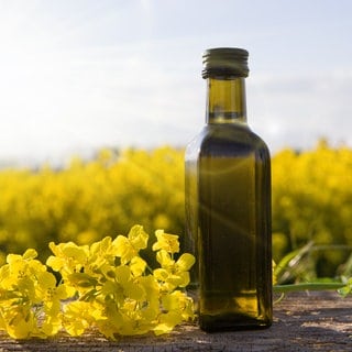 Eine flasche Rapsöl steht neben einer Rapsblüte auf einem Tisch, im Hintergrund ist ein gelb leuchtendes Rapsfeld zu sehen.