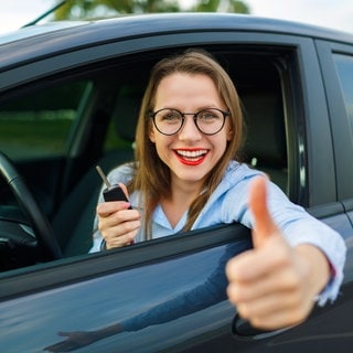 Junge Frau schaut aus Autofenster und hält stolz Autoschlüssel vor sich. Tipps und Tricks für den Gebrauchtwagenkauf. 