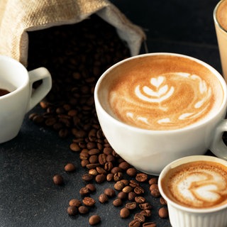 Cappucchino und Espresso stehen auf dunkler Platte, auf der Kaffeebohnen liegen. Warum ist Kaffe so teuer? Steigen die Kaffeepreise 2025 weiter?