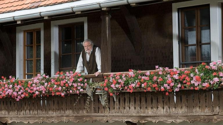 Hermann schaut vom Balkon herunter, an dem üppig die Geranien hängen