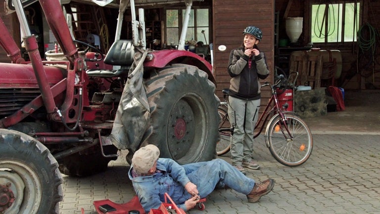 Sophie steht bei Karl, der gerade am Traktor schraubt 