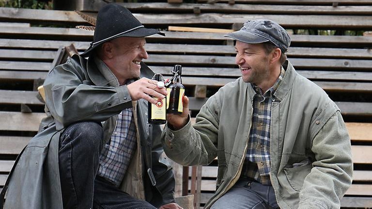 Toni und Niki sitzen vor dem Sägewerk und stoßen mit Bier an