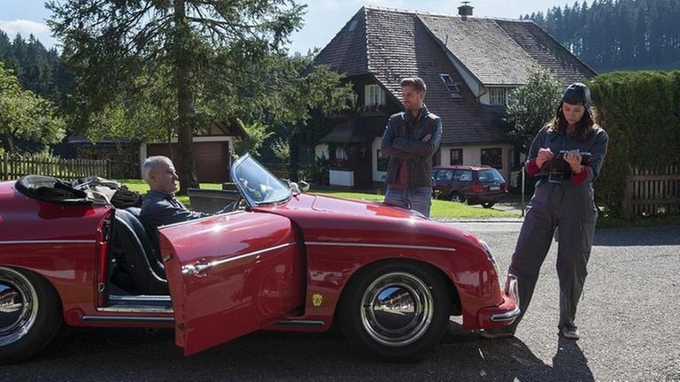 Karl sitzt in Sabines rotem Oldtimer, Andreas und Sabine stehen dabei