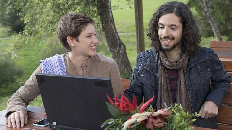 Celine und Albert sitzen vor einem Laptop in der Außenwirtschaft des Hofladens