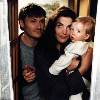 Karl und Monique, die Baby Albert auf dem Arm hat, schauen zum Fenster raus