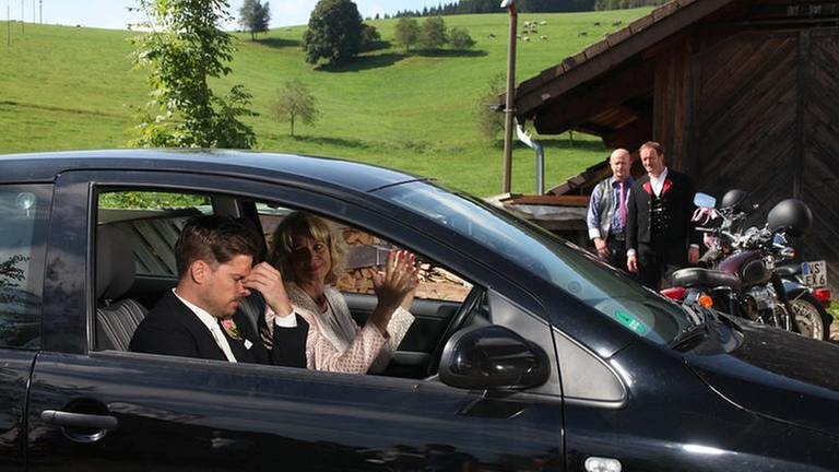 Kati sitzt mit Andreas im Auto, Bernd und Toni stehen neben ihren Motorrädern vor einer Holzhütte