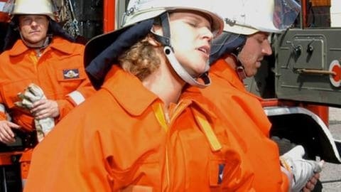 Bea und Tom sitzen in Feuerwehruniform vor einem Feuerwehrauto