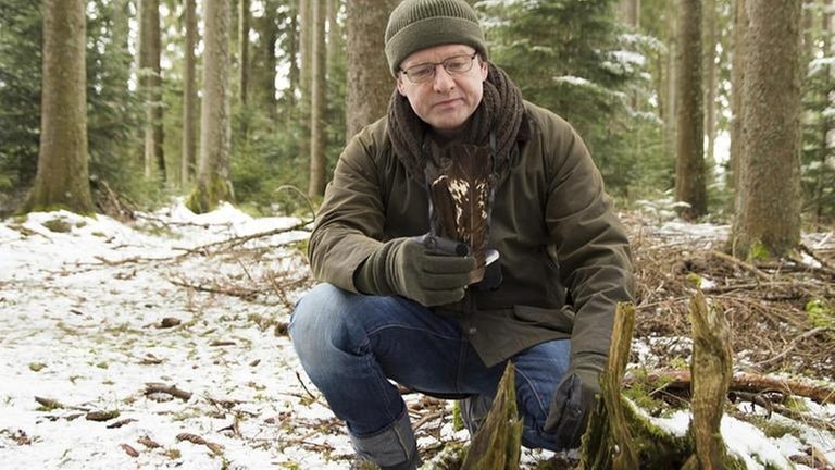 Bernhard im Wald. Er hockt vor einem Baum und hält Federn in der Hand.